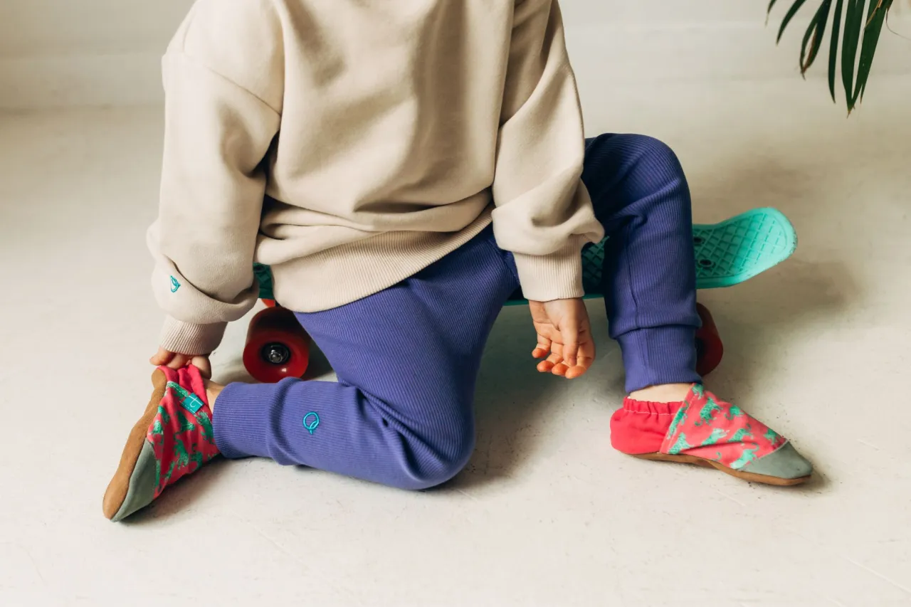 Dziecko w kolorowych kapciach, idealnych do żłobka i na fizjoterapię, siedzi na deskorolce.