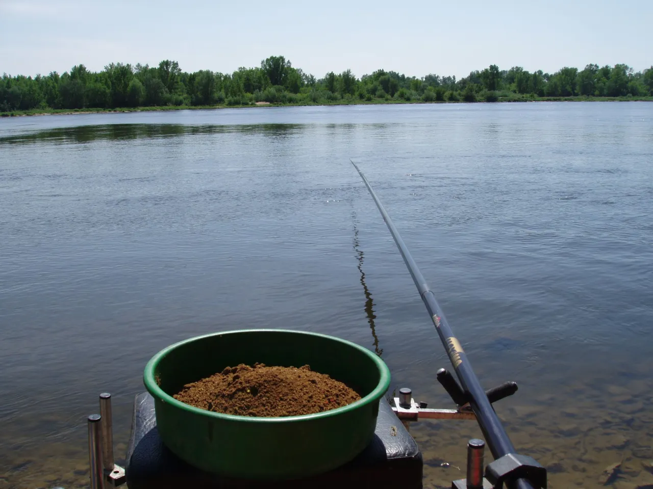 Zielona miska z sypką zanętą, idealna na płoć, czeka nad spokojną rzeką. Wędka uniesiona, gotowa do akcji.