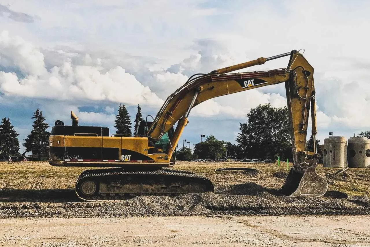 Żółta koparka CAT pracuje przy wykopie pod fundament. W tle drzewa i chmury.
