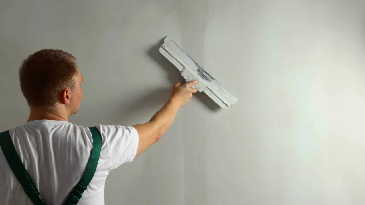 Mężczyzna w białej koszulce i zielonych szelkach nakłada gładź na ścianę z karton gips.