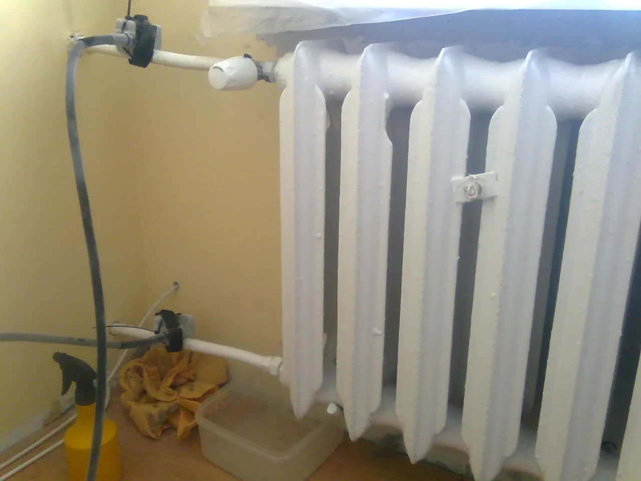 Przygotowania do tego, jak wymienić grzejnik w sezonie grzewczym: odłączanie rur, żółta butelka z rozpylaczem i pojemnik na wodę.