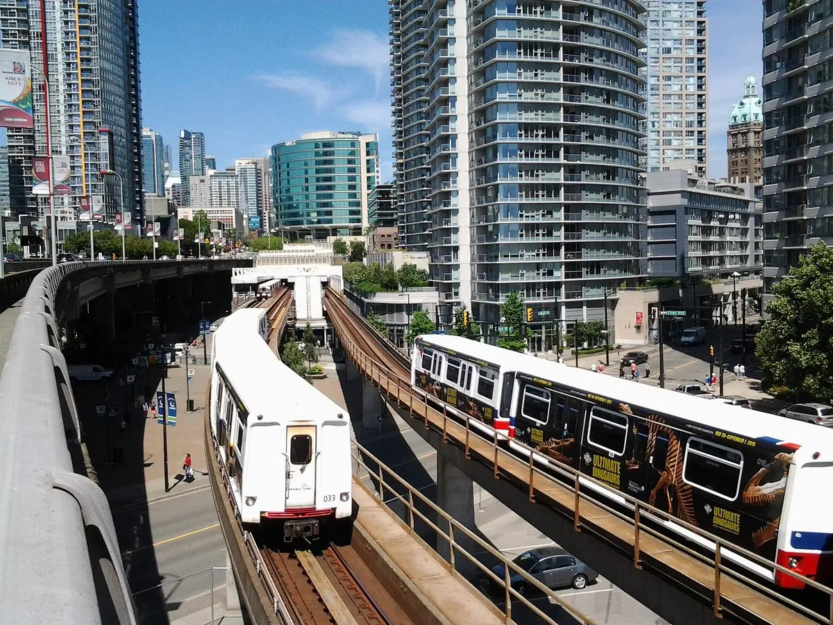 Dwa pociągi metra jadące przez nowoczesne centrum miasta, gdzie leży Vancouver. W tle wysokie budynki i błękitne niebo.