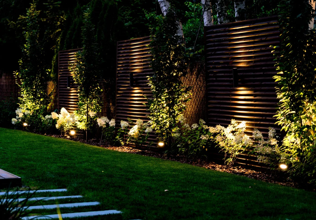 Oświetlony ogr&oacute;d przed domem z trawnikiem, krzewami i drewnianym płotem. Inspiracja, jak zaprojektować ogr&oacute;d przed domem.