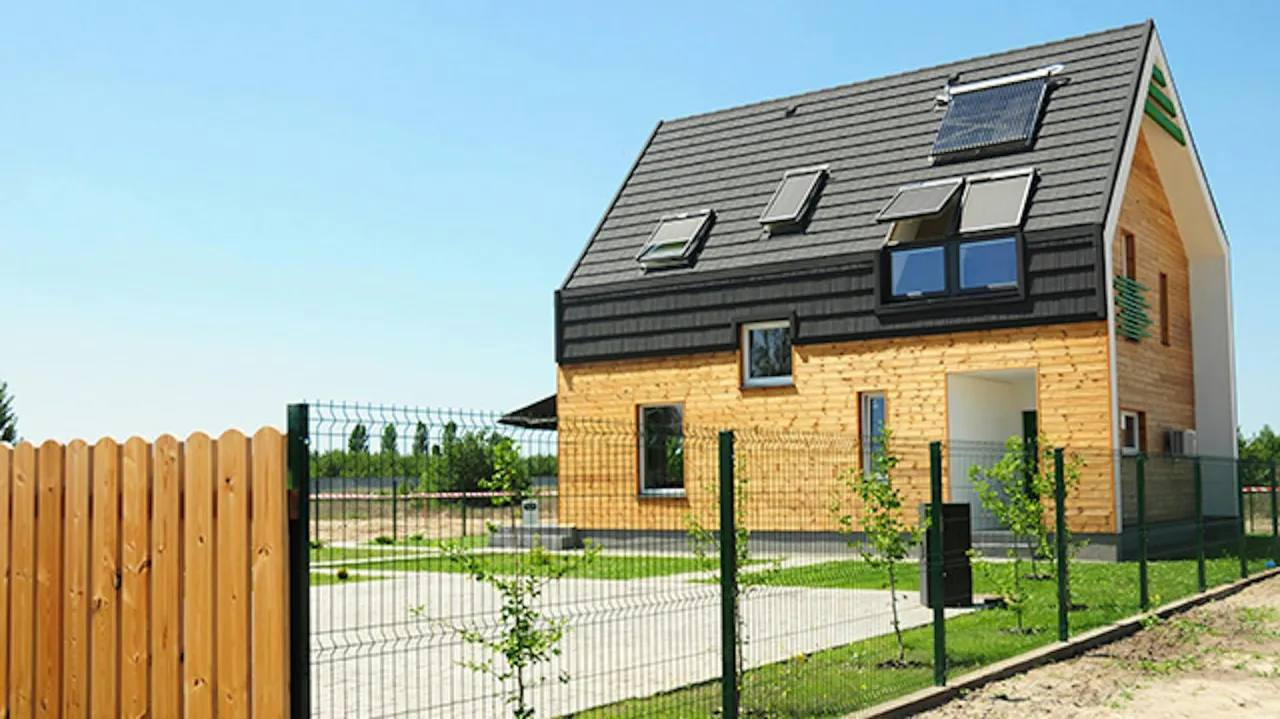 Nowoczesny, ekologiczny dom z panelami słonecznymi na dachu i drewnianą elewacją. Otoczony zielenią i ogrodzeniem.