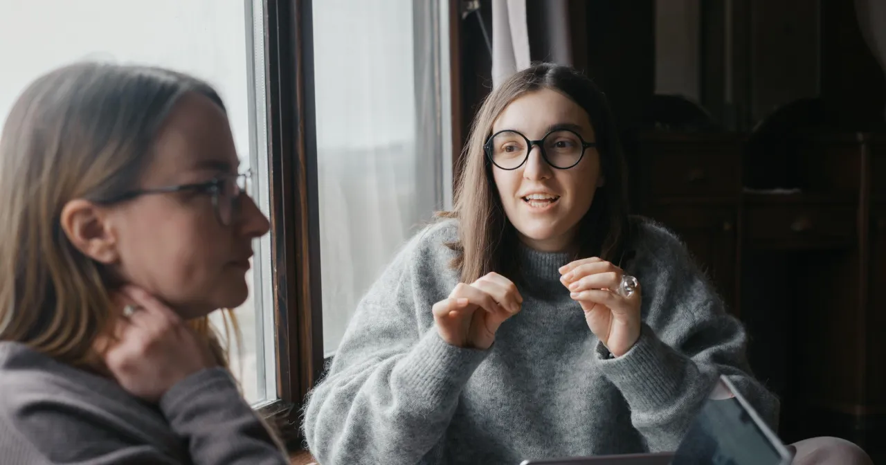 Dwie kobiety rozmawiają przy oknie. Jedna zadaje pytania drugiej poł&oacute;wce, gestykulując.