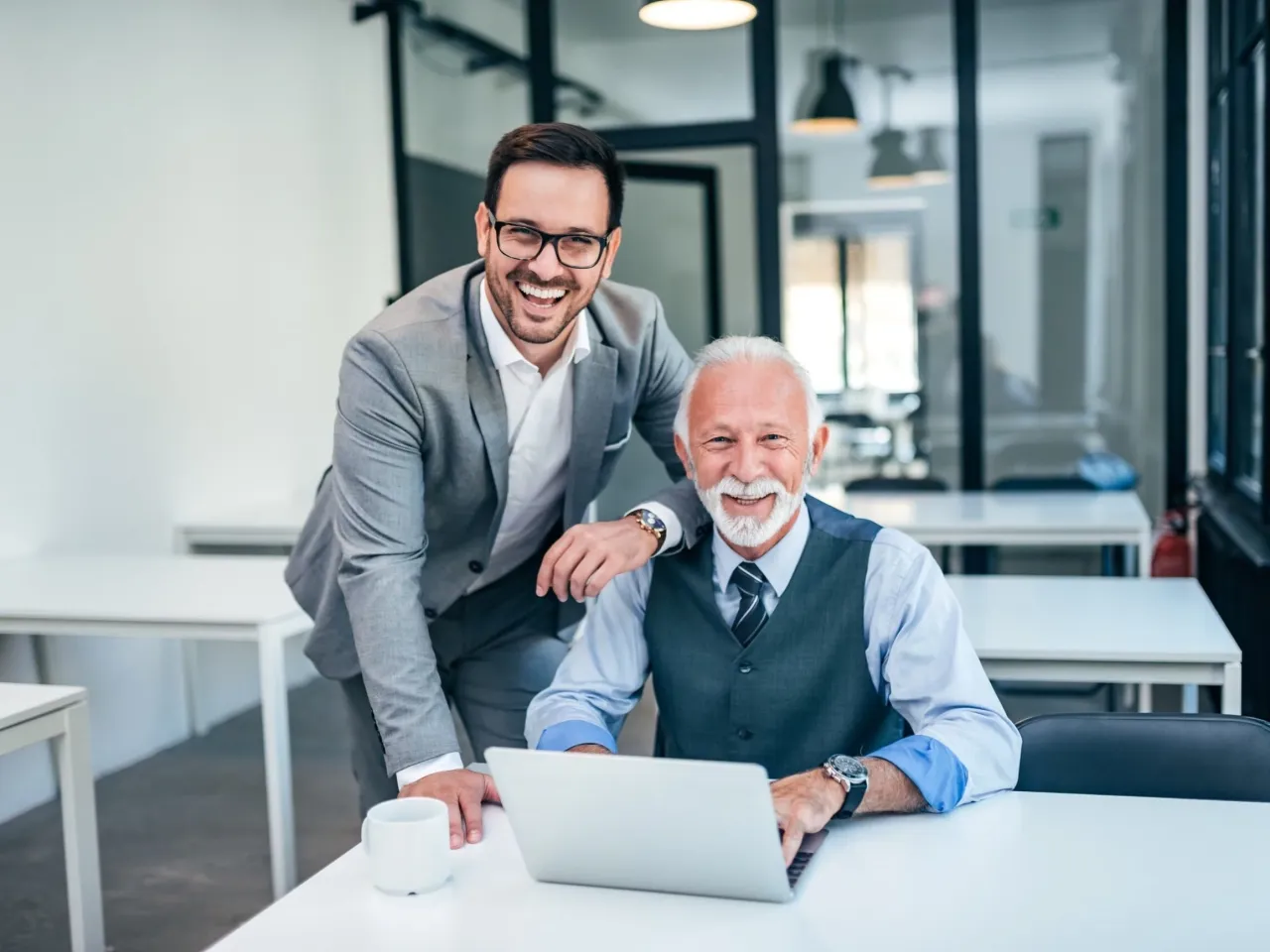 Dw&oacute;ch uśmiechniętych biznesmen&oacute;w przy biurku, jeden młodszy z okularami, drugi starszy z siwą brodą. Młodszy kładzie rękę na ramieniu starszego, pokazując mu coś na laptopie. To przykład udanej sukcesji w firmie.