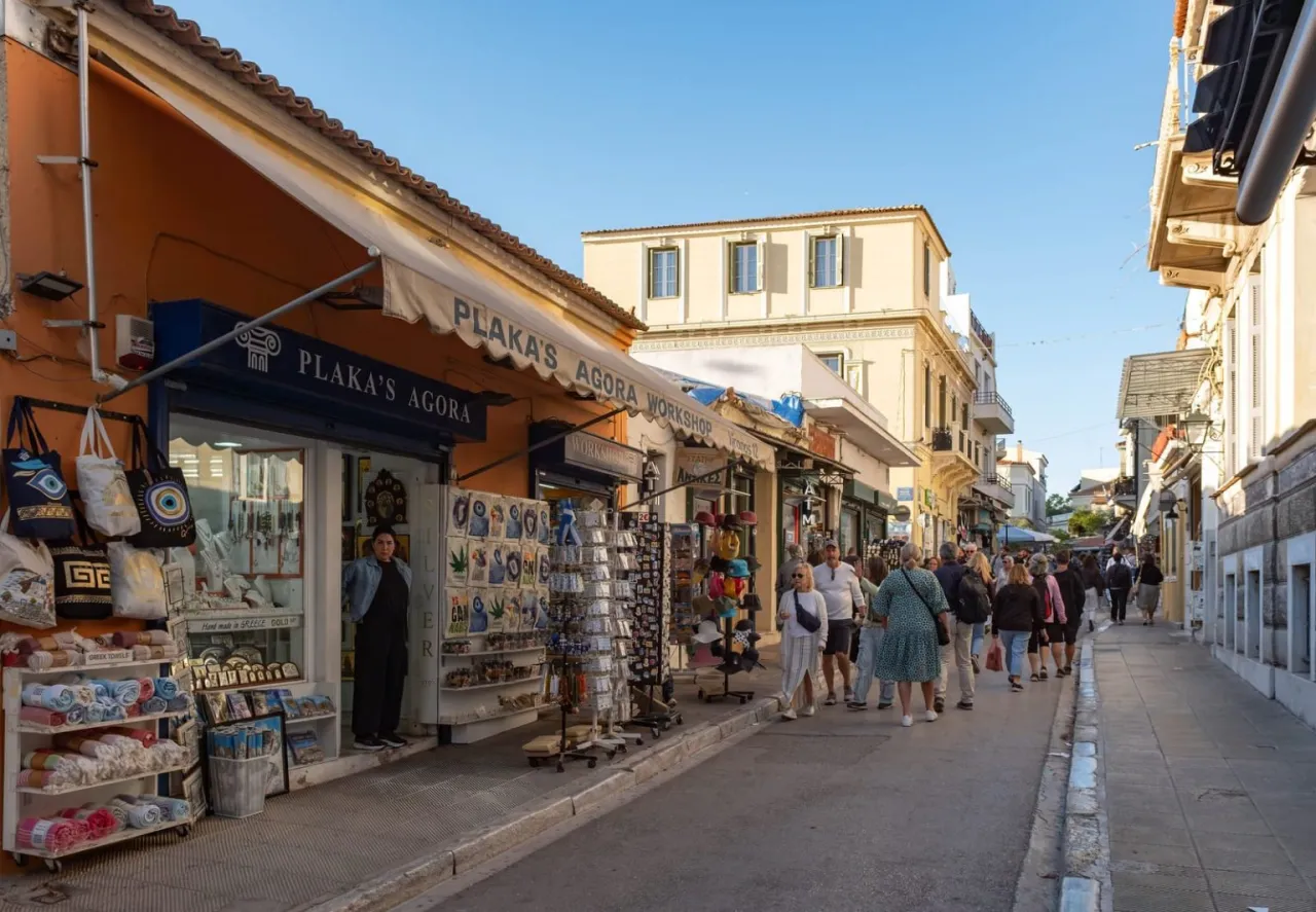 Tłum ludzi spaceruje ulicą w Atenach, gdzie sklepy z pamiątkami oferują lokalne produkty. Wygląda na to, że w Atenach jest bezpiecznie, nawet w tętniącej życiem dzielnicy Plaka.