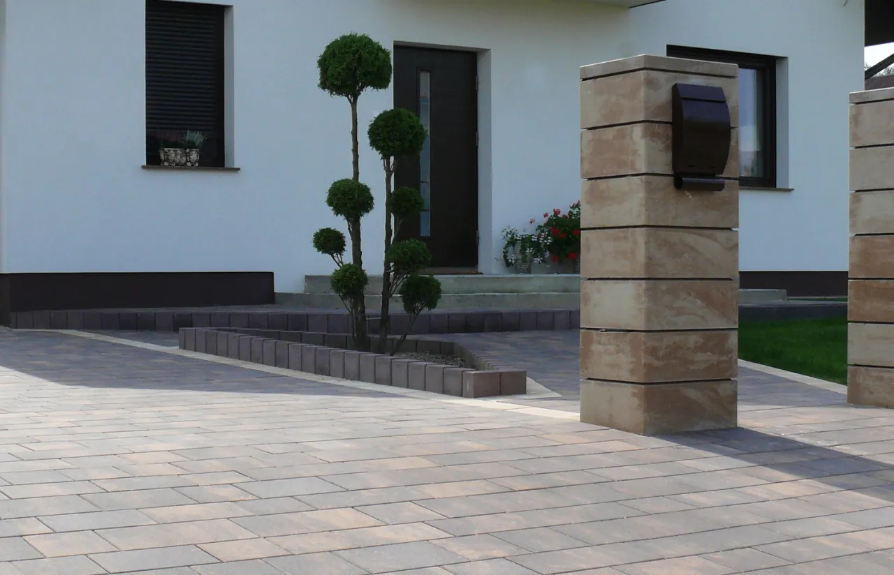 Nowoczesny podjazd z kostki brukowej, idealny jaki kamień na podjazd. Obok słupki z piaskowca i ozdobne drzewka.