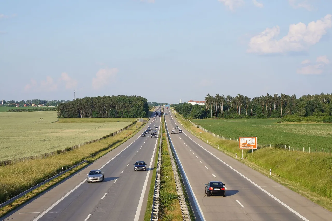 Szeroka autostrada, prawdopodobnie najdłuższa autostrada w Europie, otoczona polami i lasami. Ruch samochodowy jest umiarkowany.