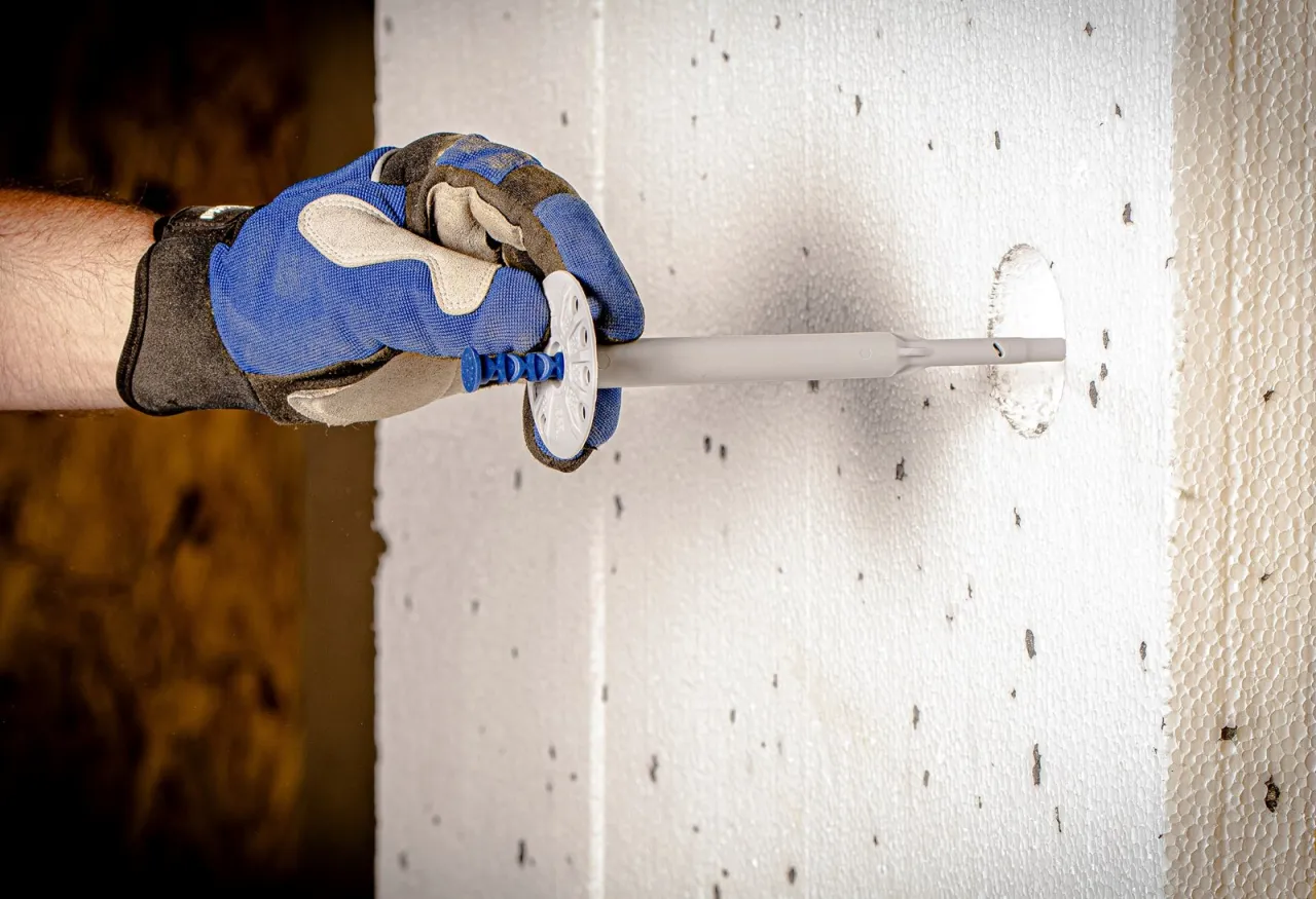 Ręka w rękawicy montuje kołki do styropianu, mocując je do białej płyty izolacyjnej.