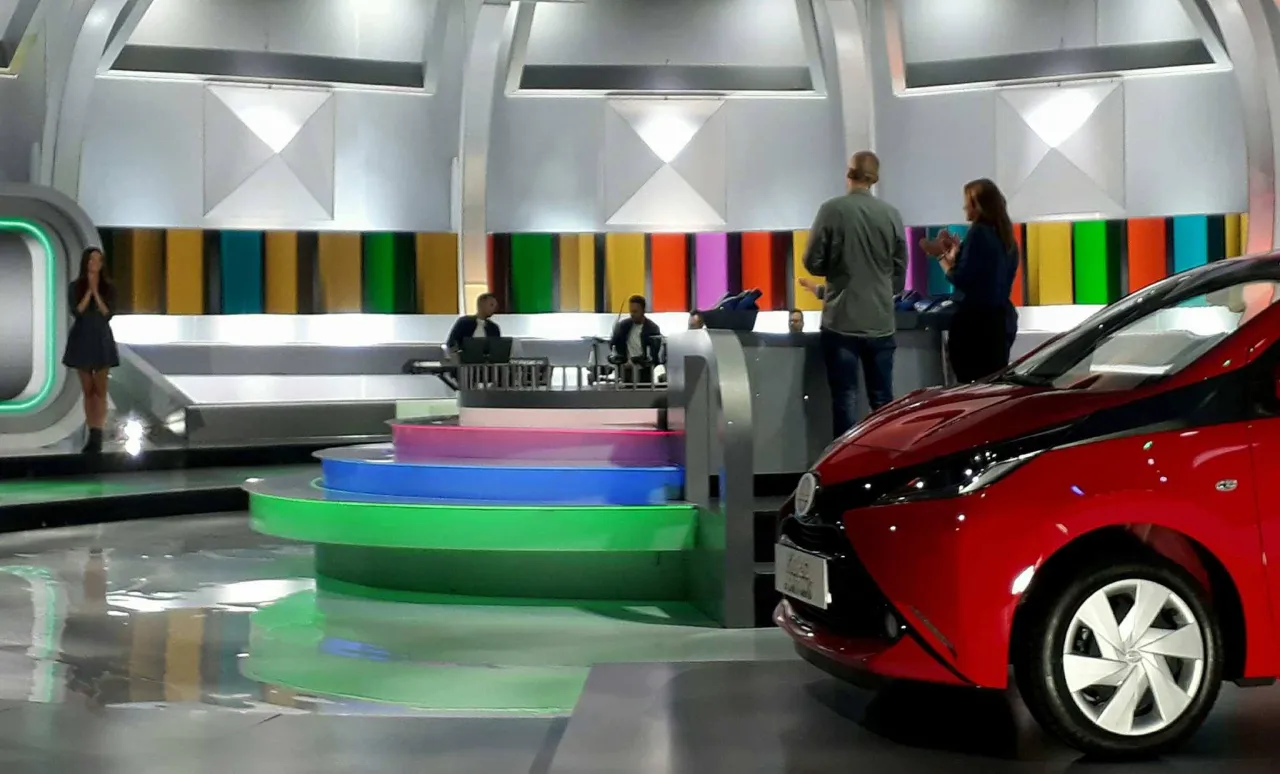Na planie programu telewizyjnego, przypominającego koło fortuny, trwają nagrania. W tle kolorowe panele, a na pierwszym planie czerwony samoch&oacute;d.