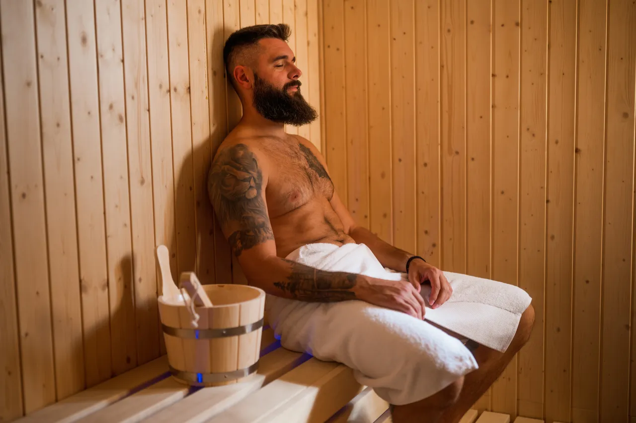 Mężczyzna z tatuażami odpoczywa w saunie. Zastanawia się, ile dni po zrobieniu tatuażu można ćwiczyć, by nie zaszkodzić gojeniu.