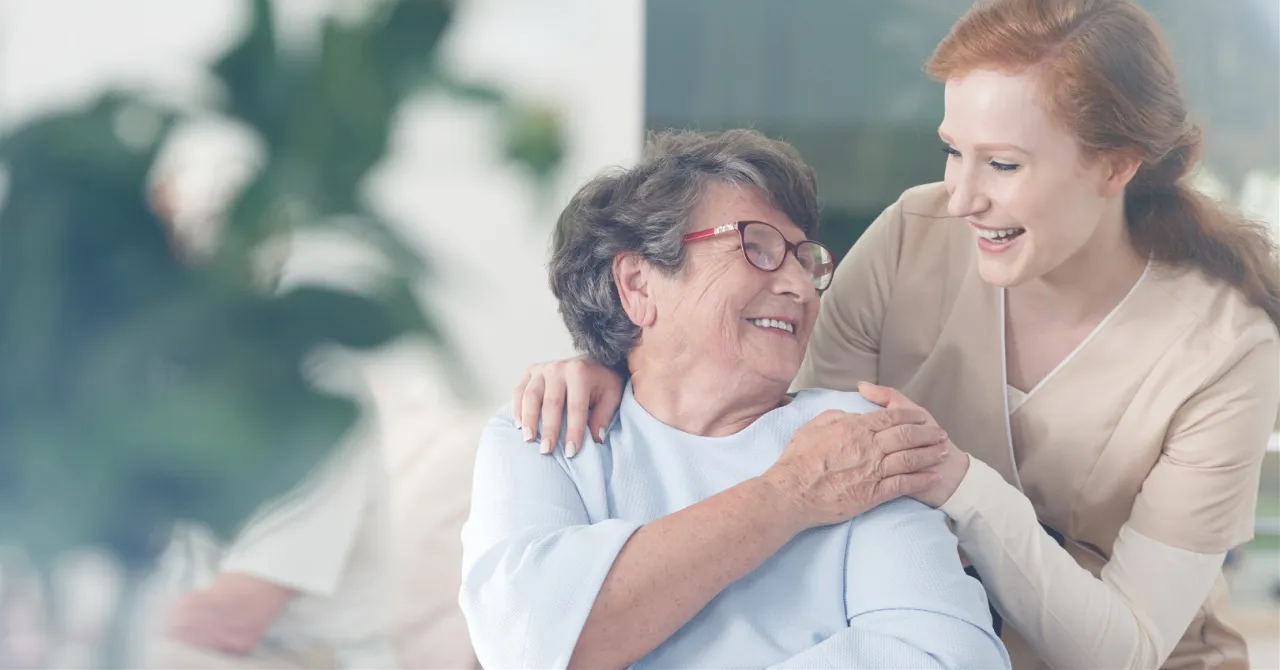 Uśmiechnięta opiekunka wspiera starszą panią. Zastanawiasz się, ile kosztuje opiekunka do starszej osoby?