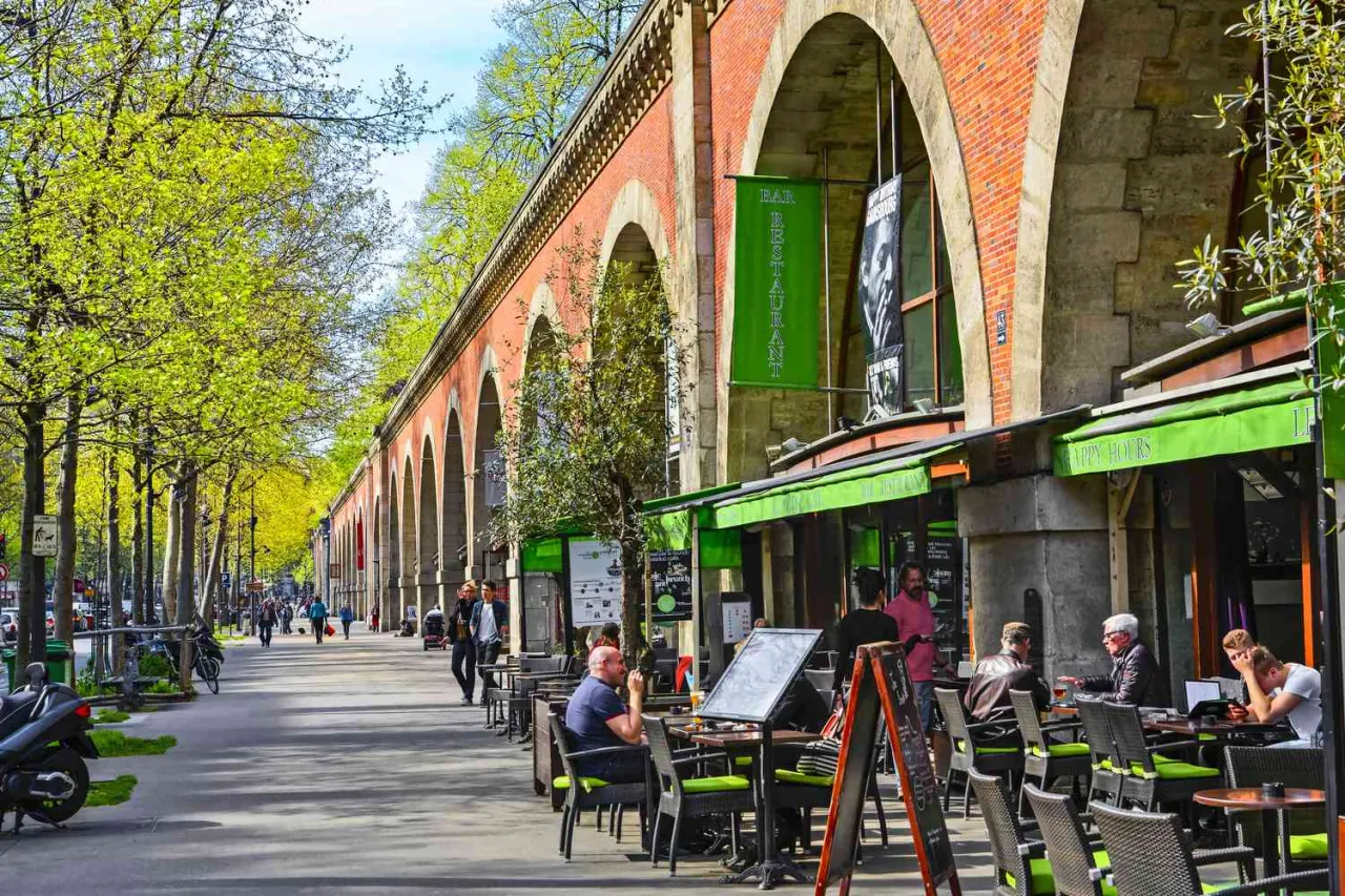 Promenada w 12 dzielnicy Paryża pod ceglanymi arkadami. Ludzie siedzą w kawiarniach, drzewa w pełni zieleni.