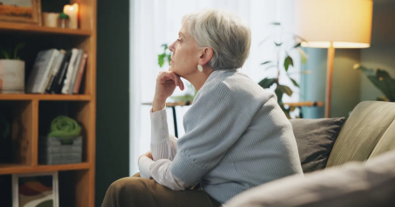 Starsza pani siedzi na kanapie, zamyślona, zastanawia się, jak odejść od męża. W tle regał z książkami i lampa.
