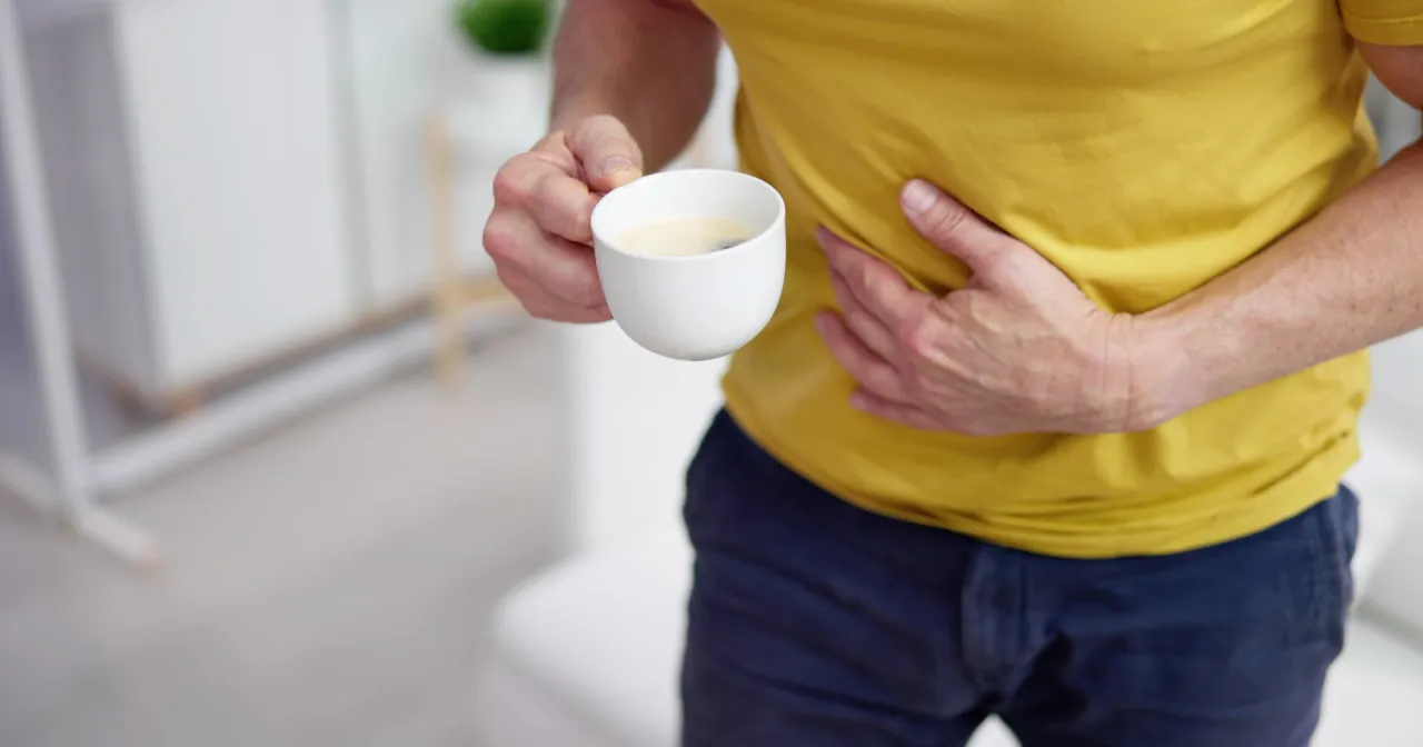 Mężczyzna trzymający kubek z kawą, masujący bolący brzuch. Szuka czegoś, co pomaga na b&oacute;l brzucha.