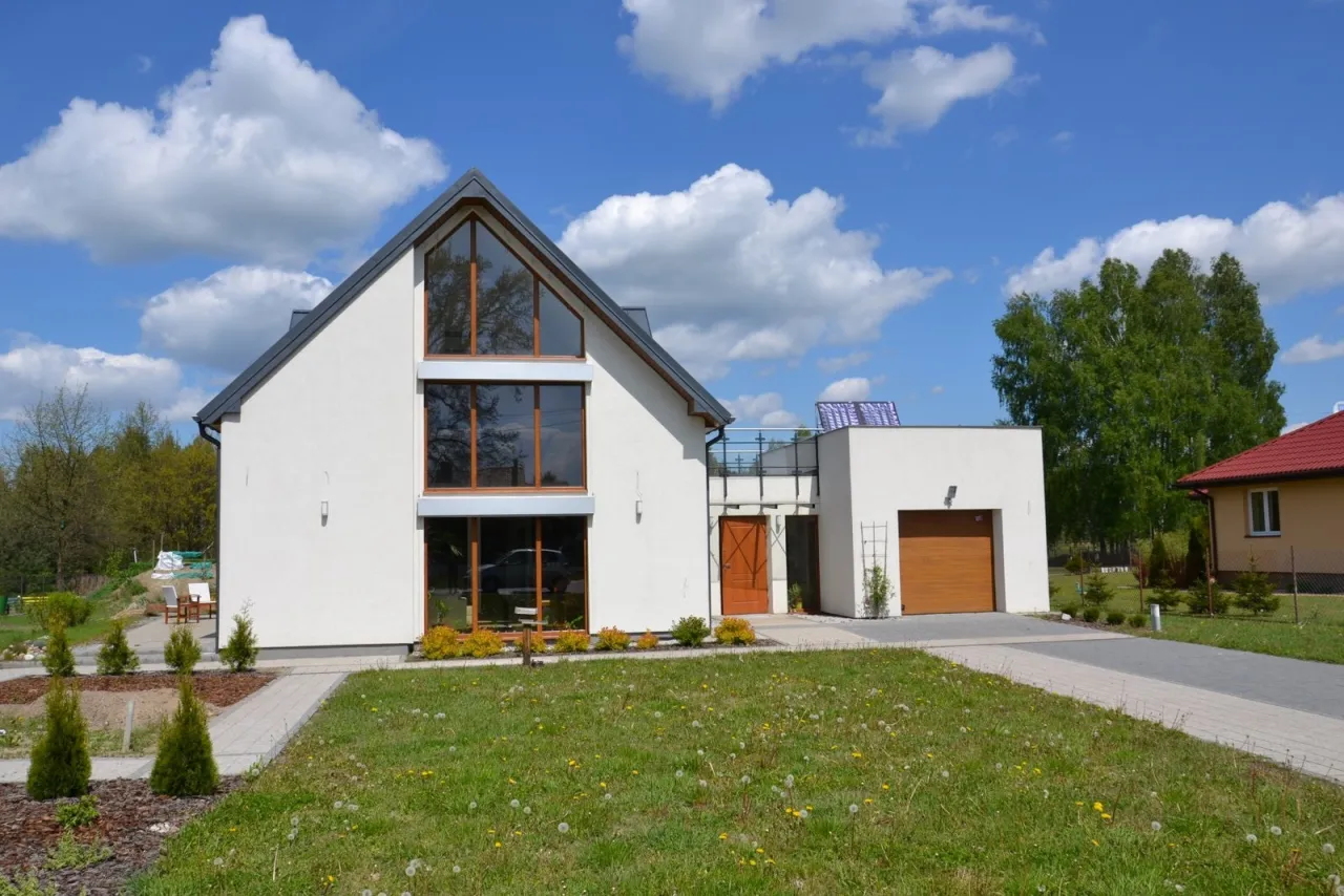 Nowoczesny dom z białym, gładkim tynkiem, który wygląda jak akryl do elewacji. Duże okna, drewniana brama garażowa i zielony trawnik.