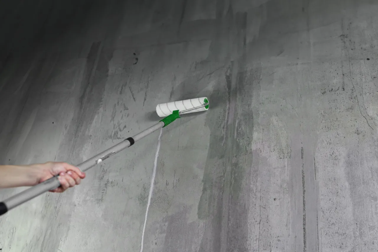 Gruntowanie ścian i sufitów przed tynkowaniem. Wałek z teleskopowym uchwytem rozprowadza preparat po szarej, betonowej powierzchni, przygotowując ją do dalszych prac.