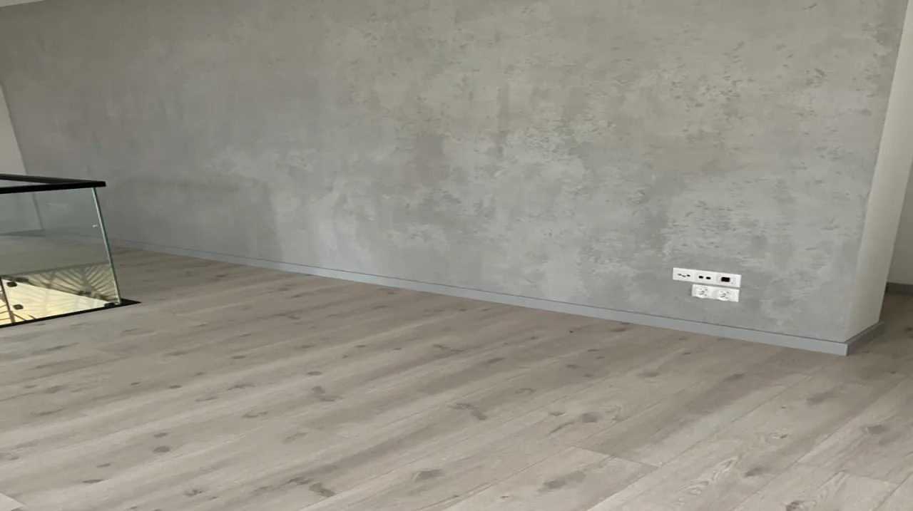 Jasna podłoga z paneli ułożona wzdłuż ściany z betonową fakturą. Widać też szklaną balustradę. Jak układać panele kierunek?