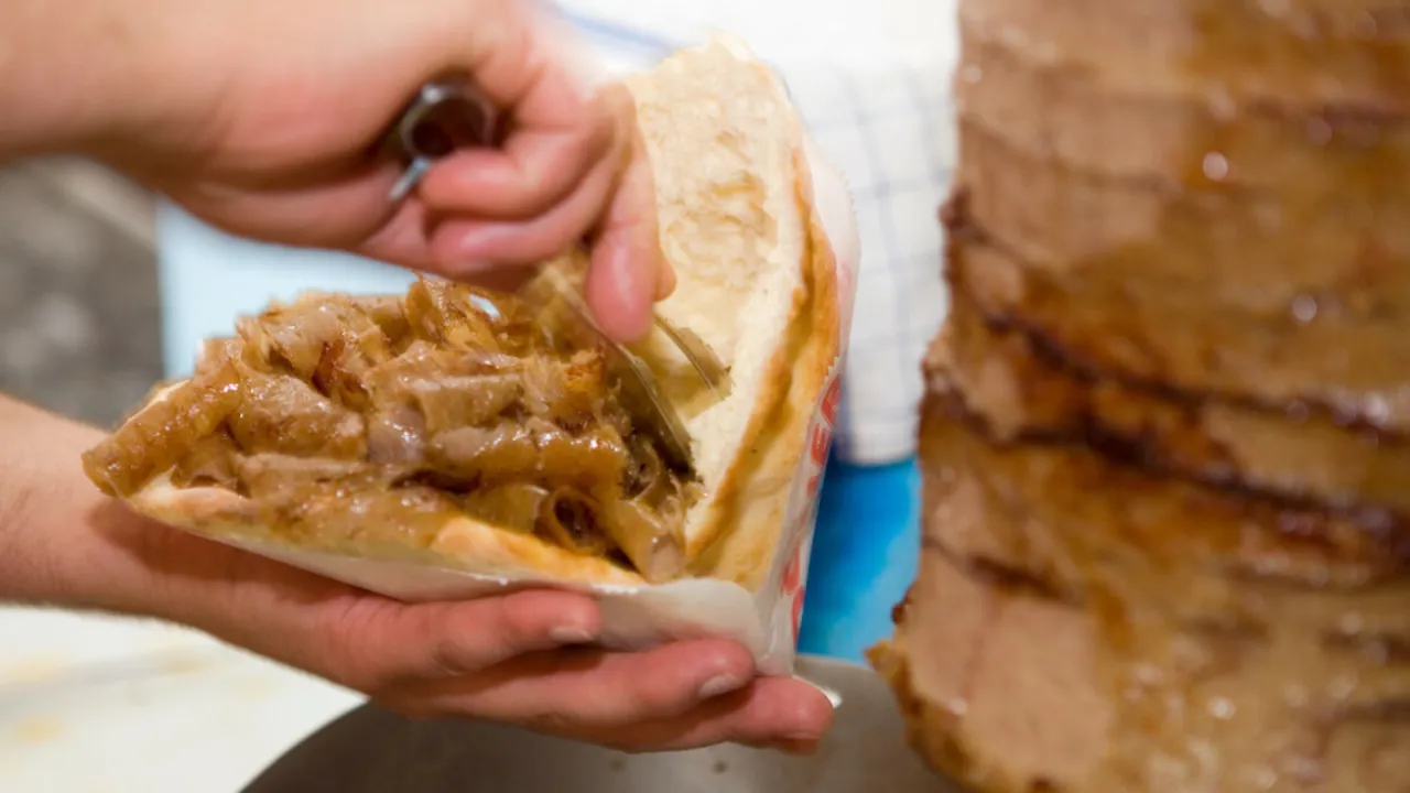 Ręce trzymają bułkę z soczystym mięsem do kebaba, obok widać pionowy rożen z pieczonym mięsem.