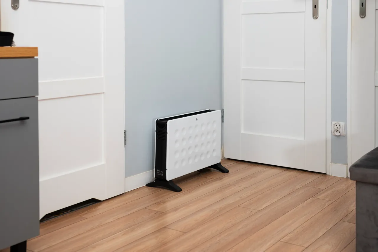 Biały grzejnik olejowy czy na podczerwień stoi na drewnianej podłodze między białymi drzwiami.