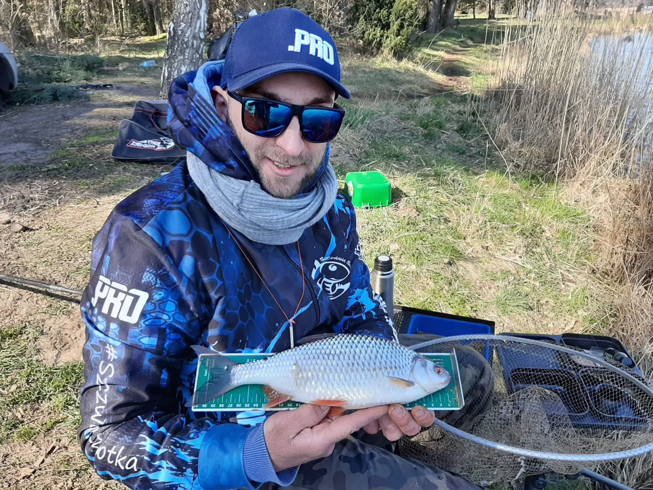 Wędkarz z rybą na miarce, w tle las i jezioro. Może to efekt stosowania receptur zanęt Górek?