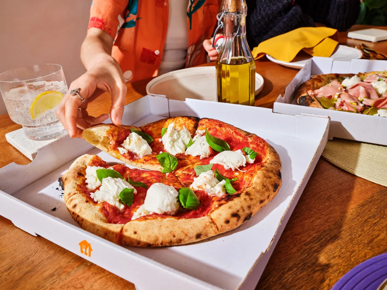 Ręka sięga po kawałek pizzy margherita, idealnej na dzień pizzy. Obok szklanka z wodą i cytryną, butelka oliwy.