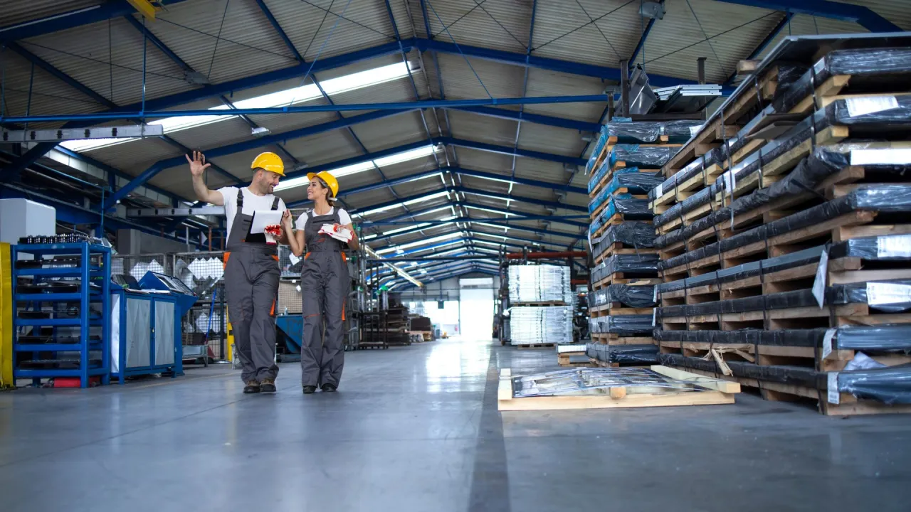 Dw&oacute;ch pracownik&oacute;w w kaskach omawia plany, jak założyć firmę budowlaną z dofinansowaniem, w magazynie pełnym materiał&oacute;w budowlanych.