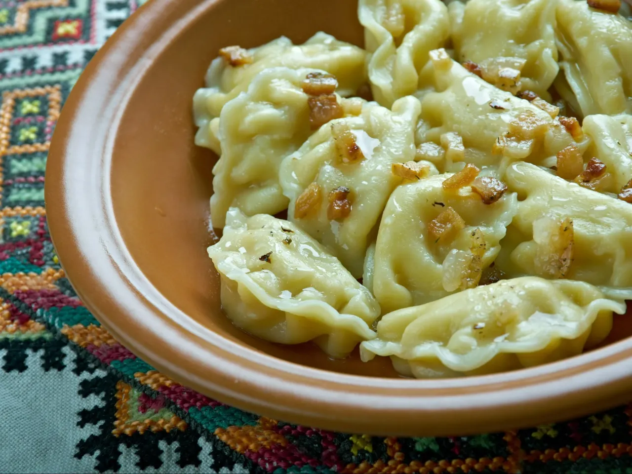 Pyszne pierogi z okrasą w brązowej misce na haftowanej serwetce. Trudno oszacować, ile sztuk pierogów wchodzi na kilogram, ale porcja wygląda na obfitą.