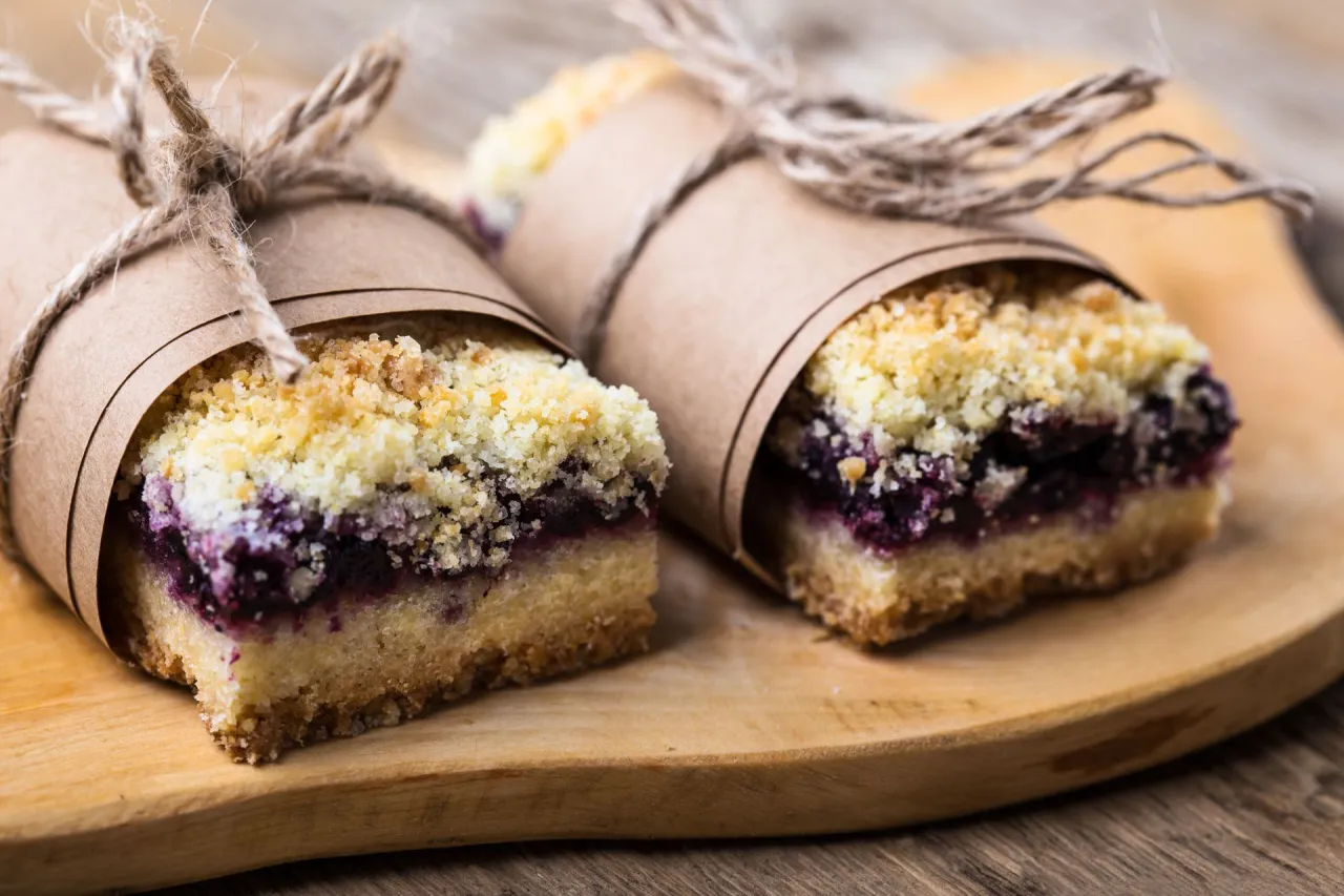 Dwa kawałki biszkoptu z jagodami i kruszonką, zawinięte w papier i przewiązane sznurkiem, leżą na drewnianej desce.