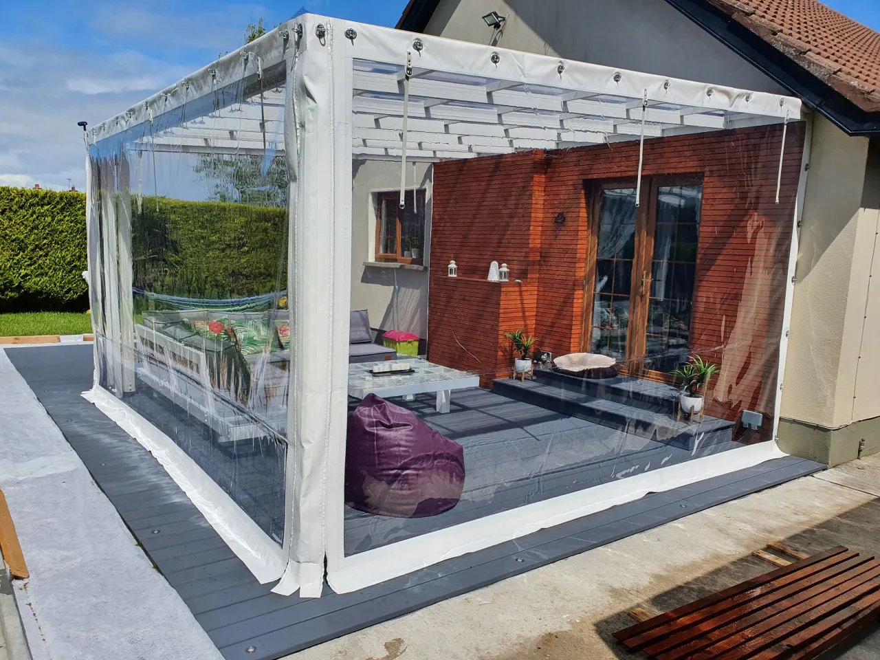 Zabezpieczona altanka z przezroczystymi ścianami chroni przed wiatrem. Wewnątrz meble wypoczynkowe i stolik.