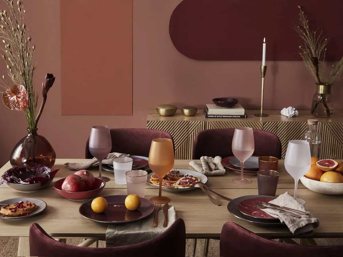 Elegancko nakryty stół, gdzie kolor burgund przeplata się z ciepłymi odcieniami brązu i różu. Widać owoce, kieliszki i zastawę stołową.
