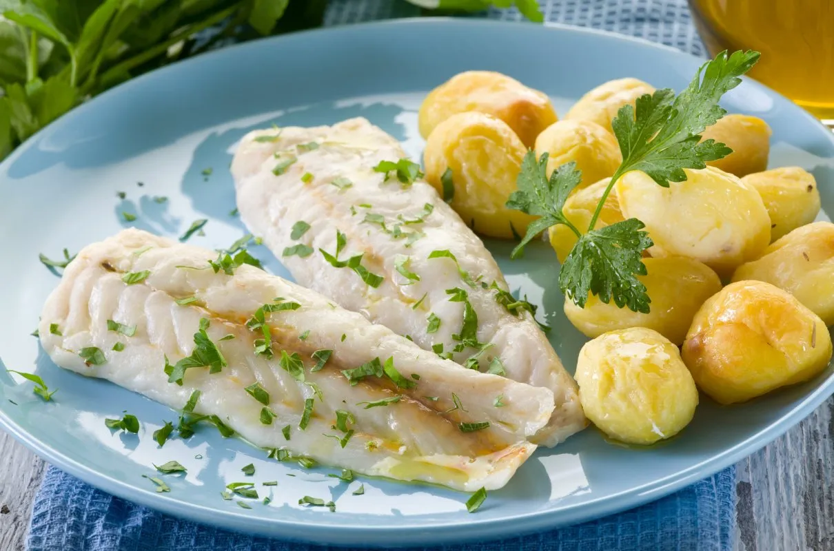 Dwa filety ryby na parze z młodymi ziemniakami i natką pietruszki na niebieskim talerzu.