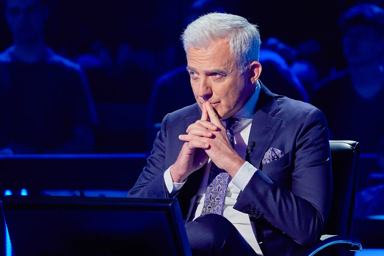 Mężczyzna w garniturze, kt&oacute;ry wolumen to wolumin milionerzy, zamyślony siedzi na tle publiczności.